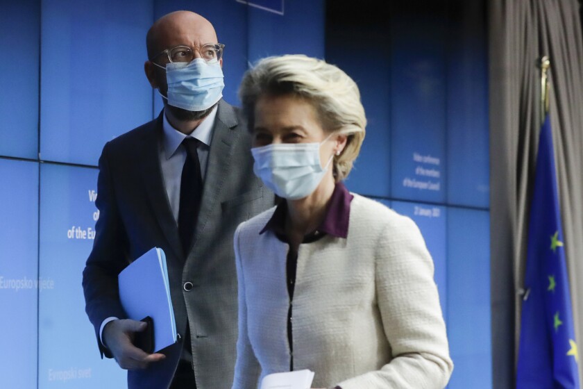 Two European government officials, in masks and carrying notes, walk across a stage.