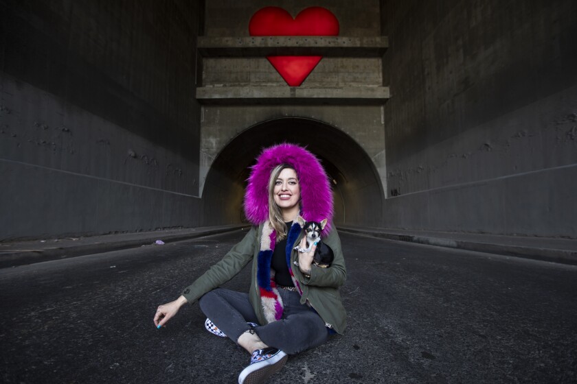 A woman sits in the road outside a tunnel, holding a chihuahua, her face framed with fuchsia faux fur.