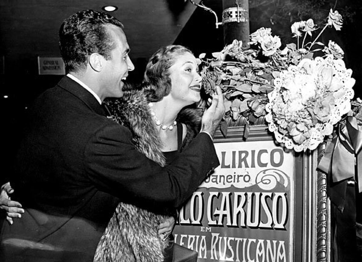 Ricardo Montalban and his wife, Georgiana, sister of Loretta Young, stop to smell the roses placed over an old Caruso poster in the lobby of the Egyptian Theatre in Hollywood at the premiere of "The Great Caruso" in 1951.