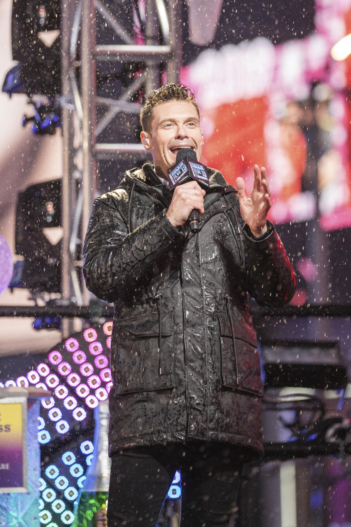 Ryan Seacrest in a black puffer jacket speaks into a microphone in the snow