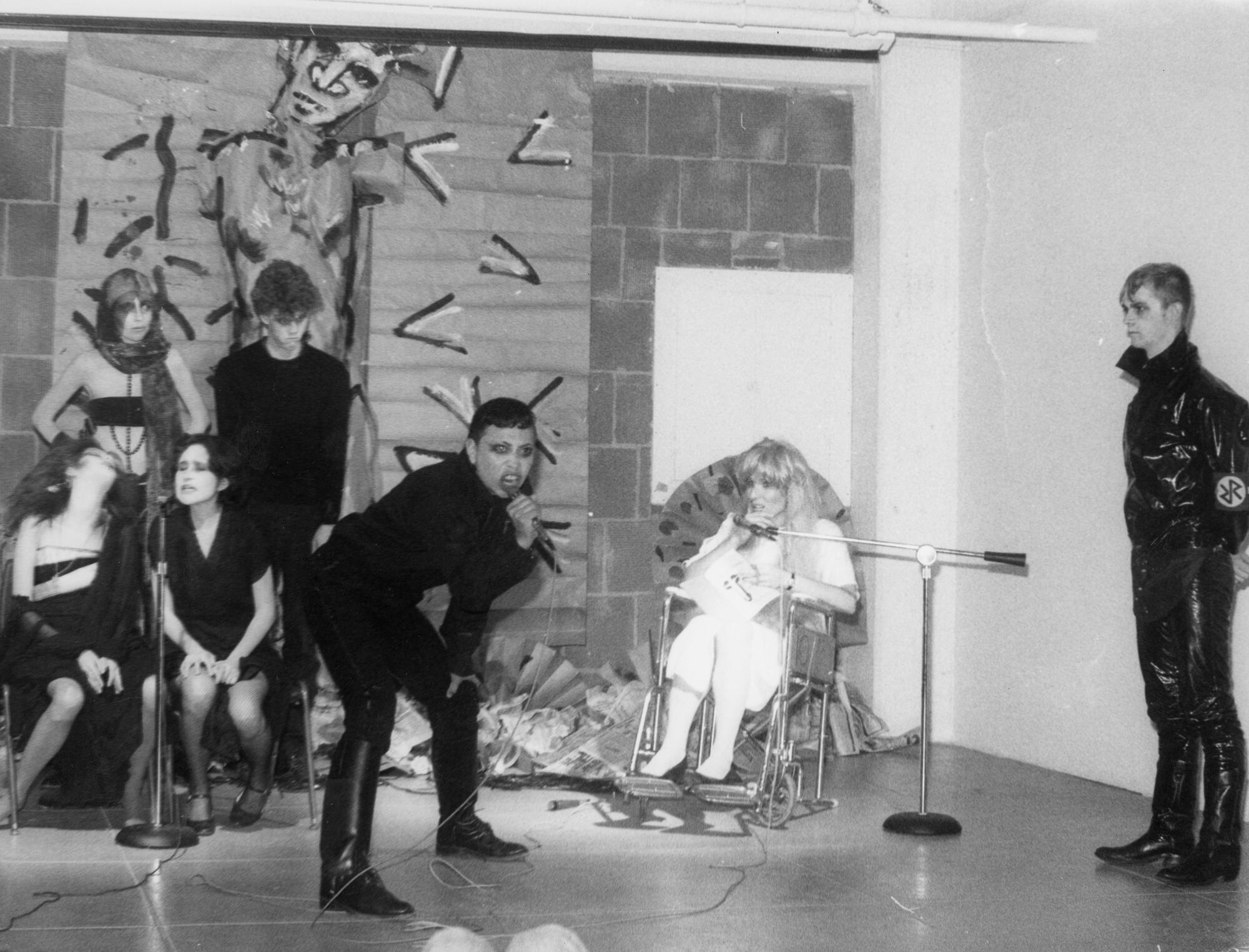 A singer dressed in black wails into a mic as various people in black and white costumes sit and stand around him.