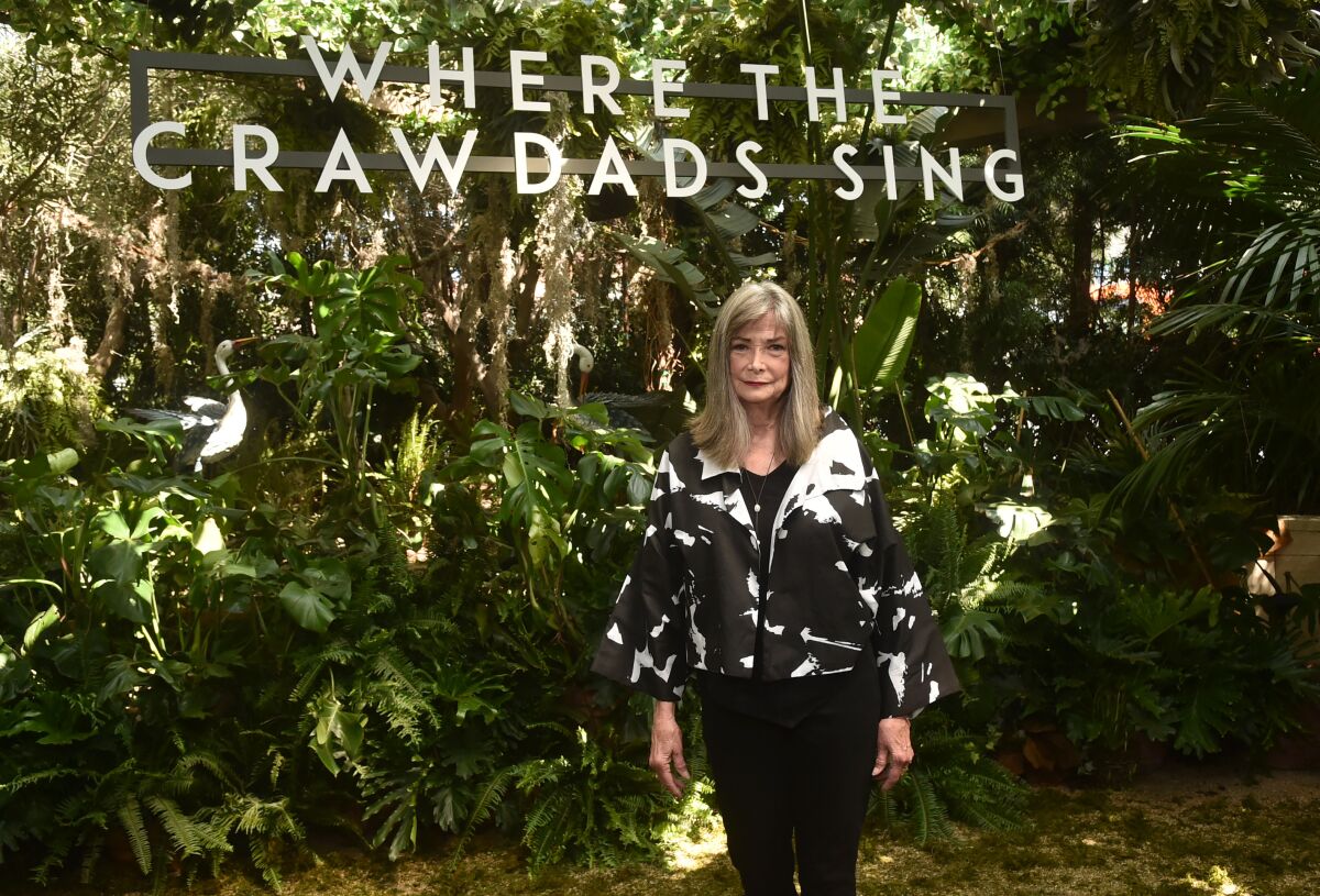 A woman dressed in black and white stands in front of greenery and a sign that reads, "Where the Crawdads Sing."