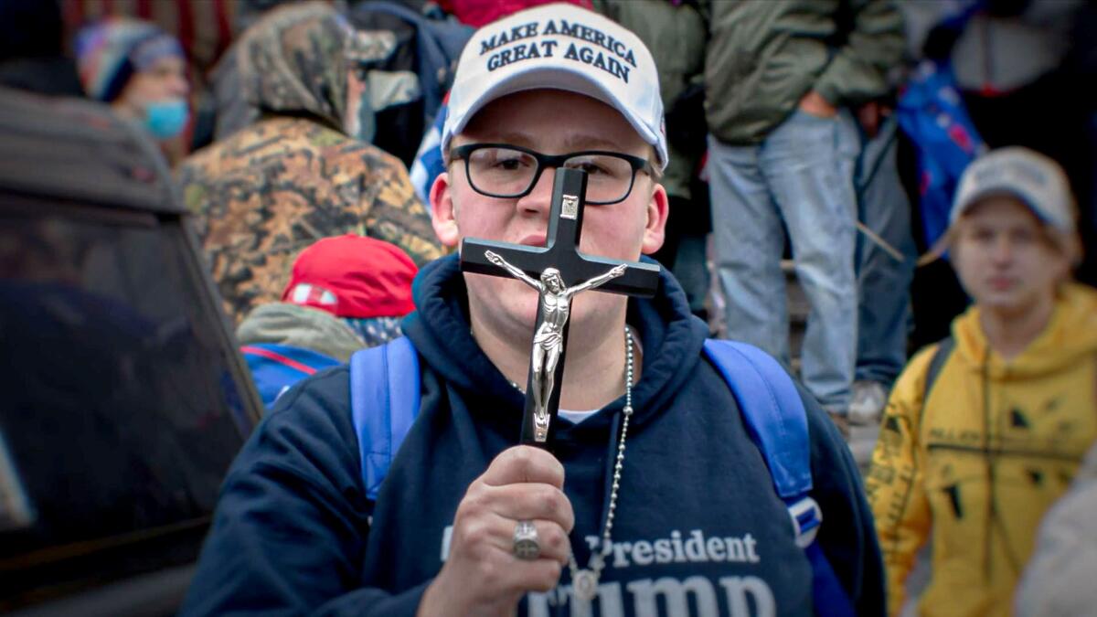 A man wearing a Trump sweatshirt and "Make America Great Again" hat holds up a crucifix.