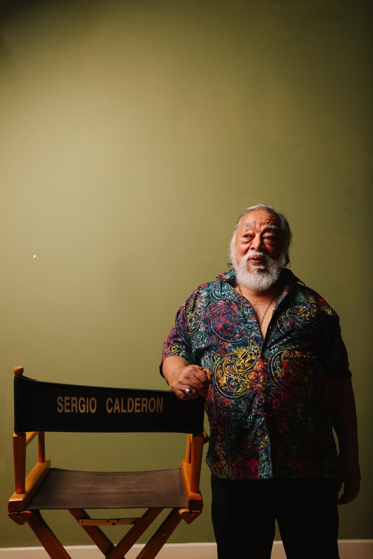 Sergio Calderón in a patterned shirt and black pants resting his right arm on a director's chair with his name on it