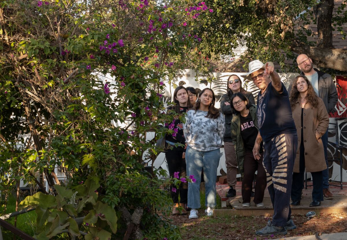 Randy King Lawrence yeşilliklerle dolu bahçesinde bir grup ziyaretçinin önünde dururken kamera dışındaki bir şeyi işaret ediyor