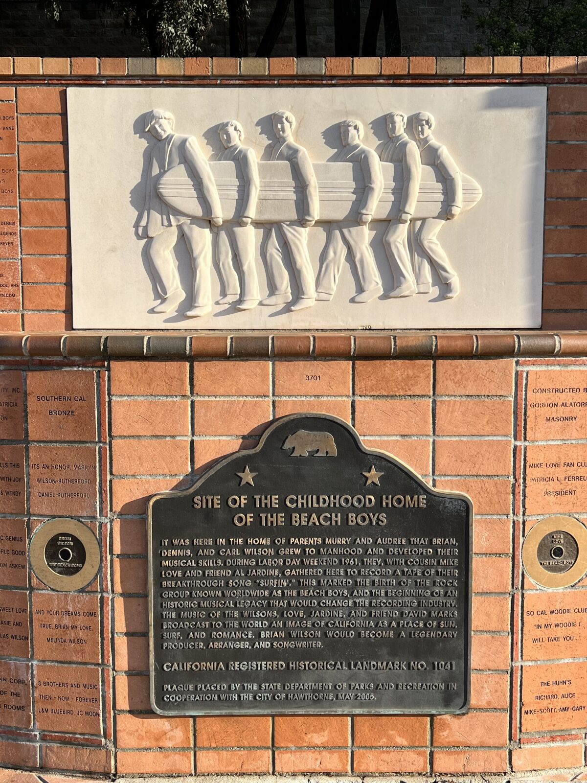 Une sculpture de six hommes tenant une planche de surf et une plaque indiquant : Site de la maison d'enfance des Beach Boys.