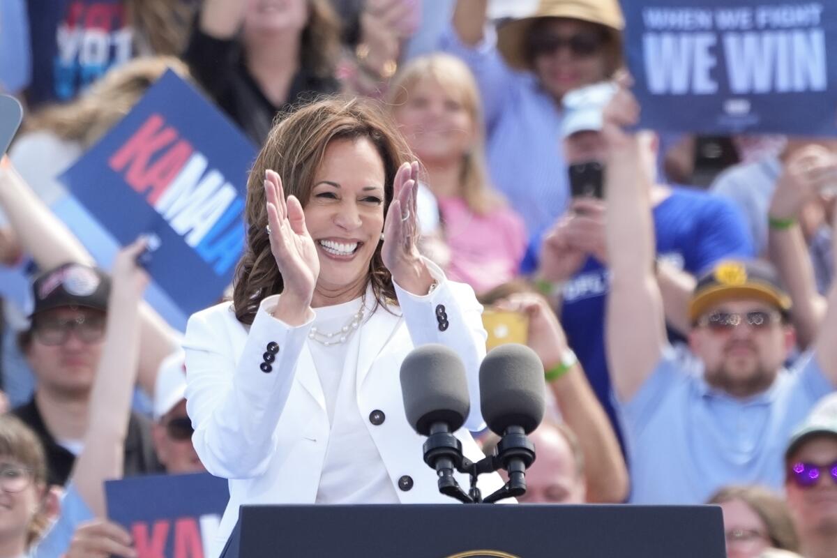 Kamala Harris smiles and claps at a rally as supporters behind her raise campaign signs, one reading "When we fight, we win"