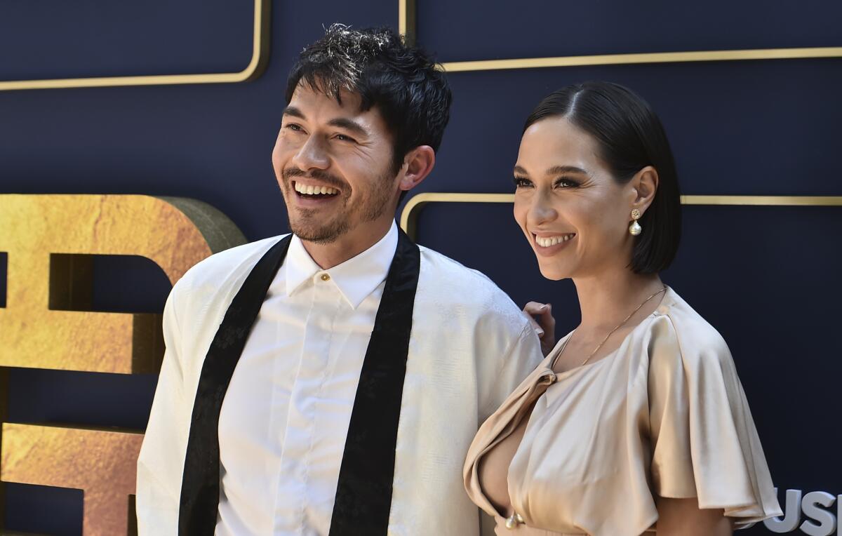 Henry Golding in a white suit jacket and shirt with a black sash, standing next to a woman in a cream colored dress