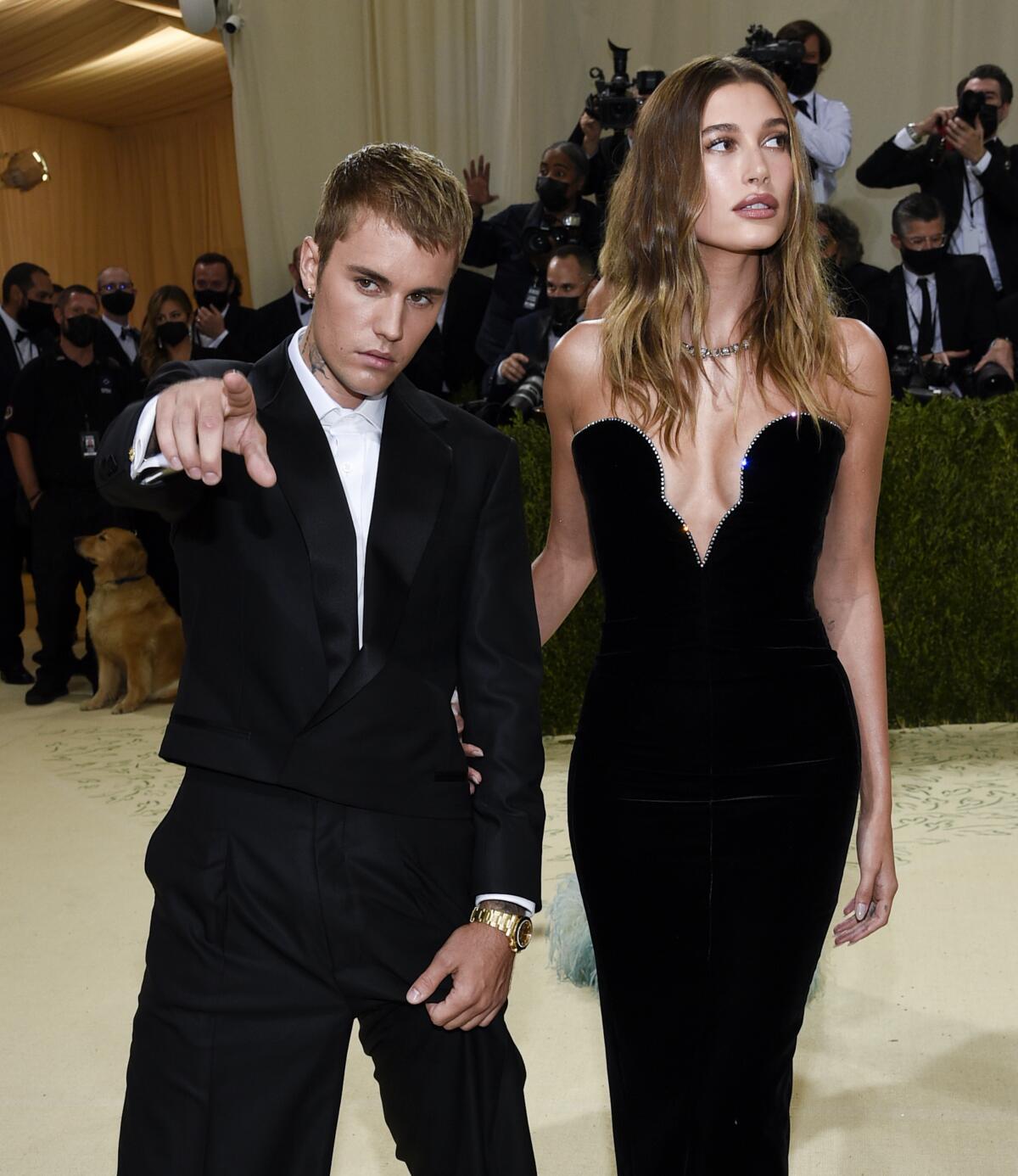 Justin Bieber in a baggy black suit points at the camera while standing next to Hailey Bieber in a black strapless gown