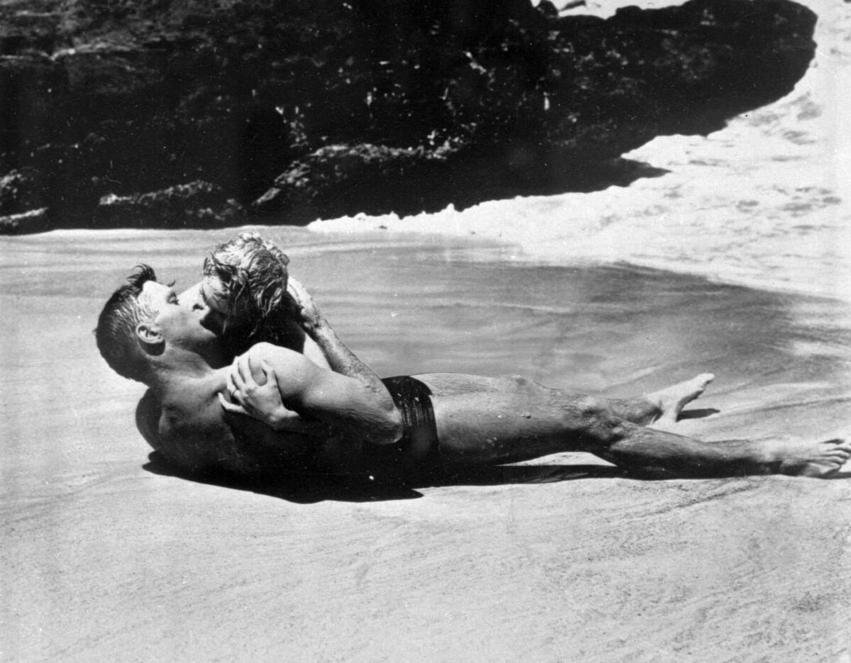 A black-and-white film still shows a couple lying on a beach kissing