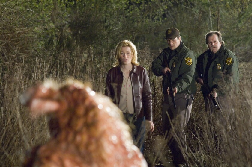 A woman and two armed law enforcement officers confront a slimy, disgusting creature in the woods at night
