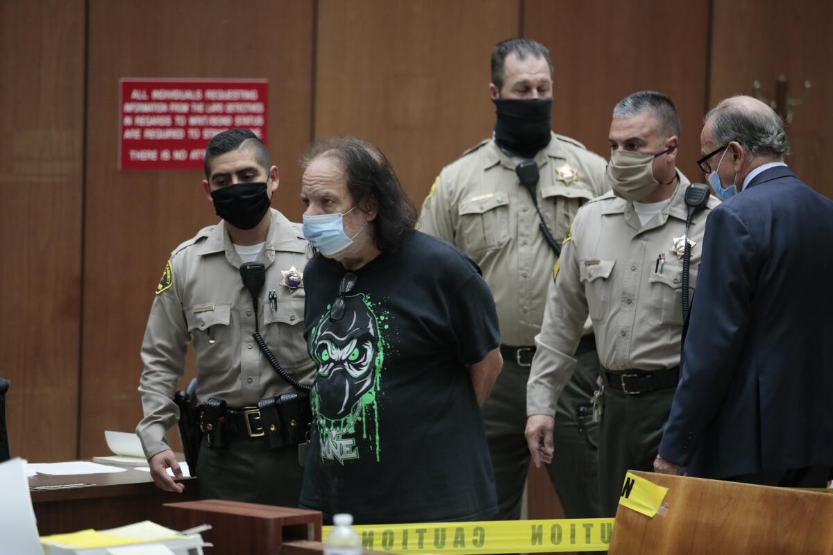 A man wearing a T-shirt and face mask stands with his hands behind his back while surrounded by law officers in a courtroom