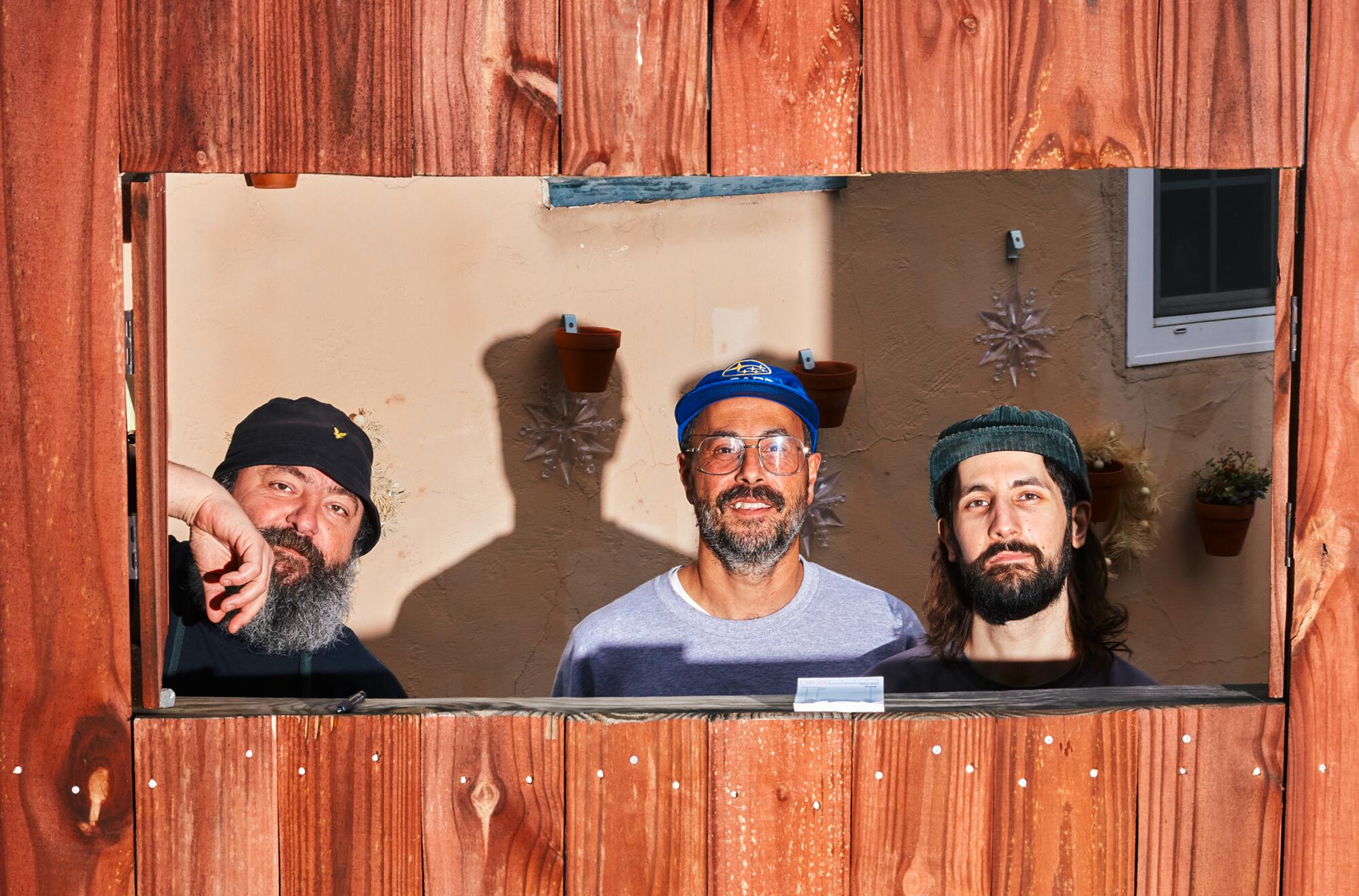 Three bearded men framed in a cutout in a wooden fence.