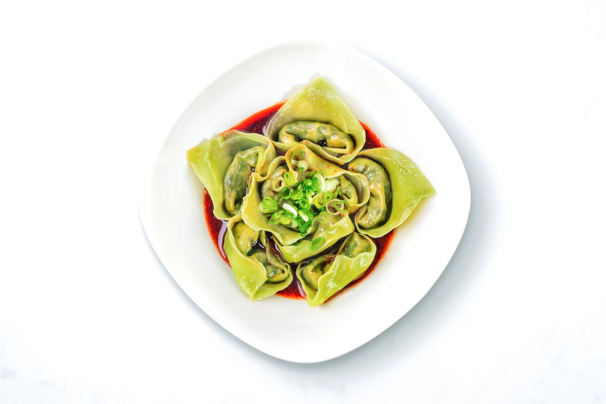 Green wontons, in a red sauce on a white plate, are arranged to resemble an opening flower.