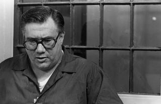 A black-and-white photo of a man wearing a dark shirt and glasses, looking down