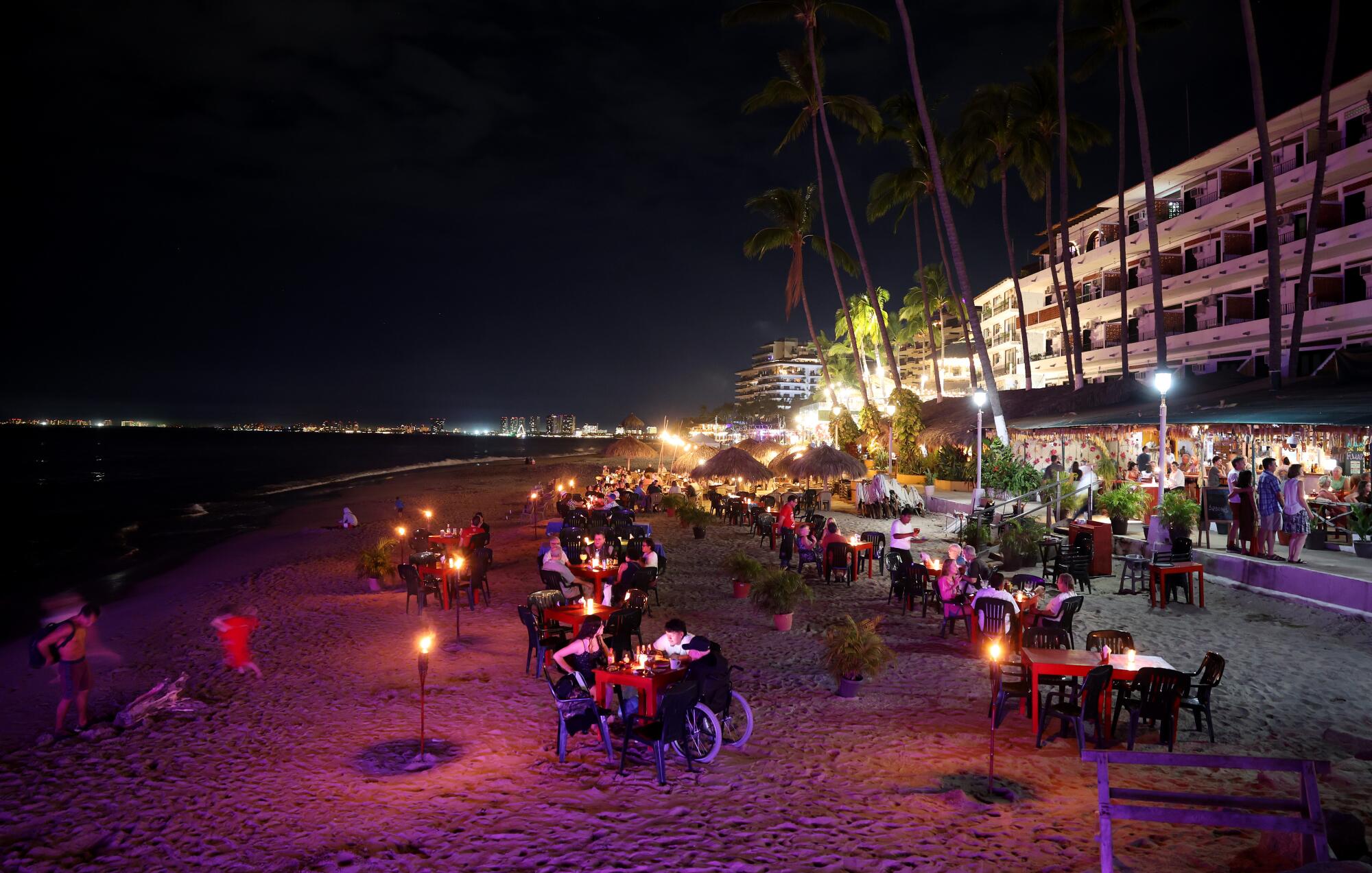 People dining on a beach under torchlight as others stroll by beachfront stores