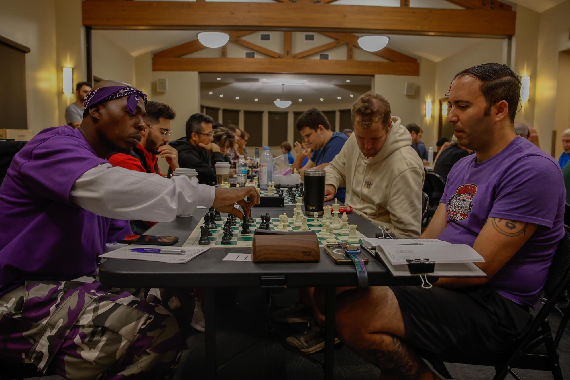 Vincent Hubbard, dressed in purple, plays chess with another man in purple at a long table of chess players at a tournament.