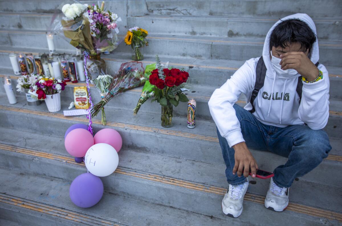 A teen boy covers his face while sitting next to flowers, candles and balloons placed on school steps