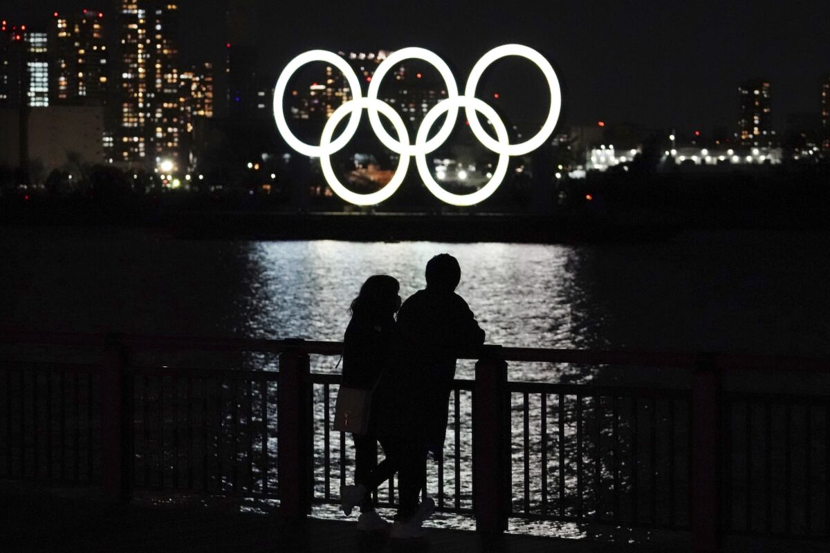 A man and a woman in silhouette on a bridge at night with the Olympic rings illuminated in the background.
