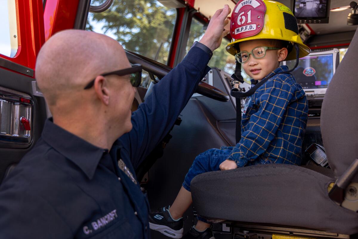 A firefighter stands next to a young boy seated in a firetruck
