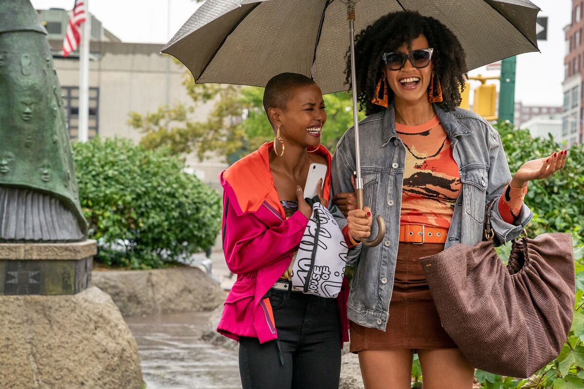 Two fashionably dressed women laugh together under an umbrella as they walk on a city sidewalk.