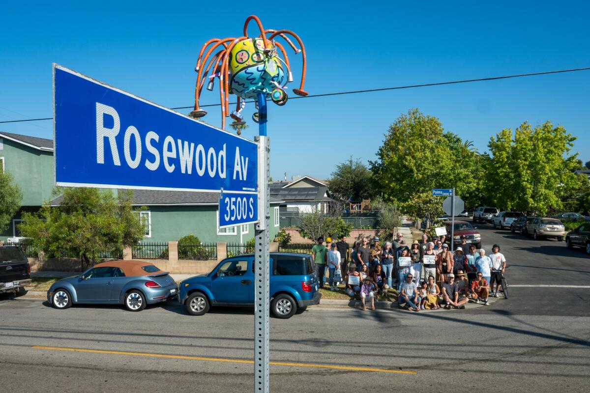 A street sign topped with a sculpture and people gathered across the street. 