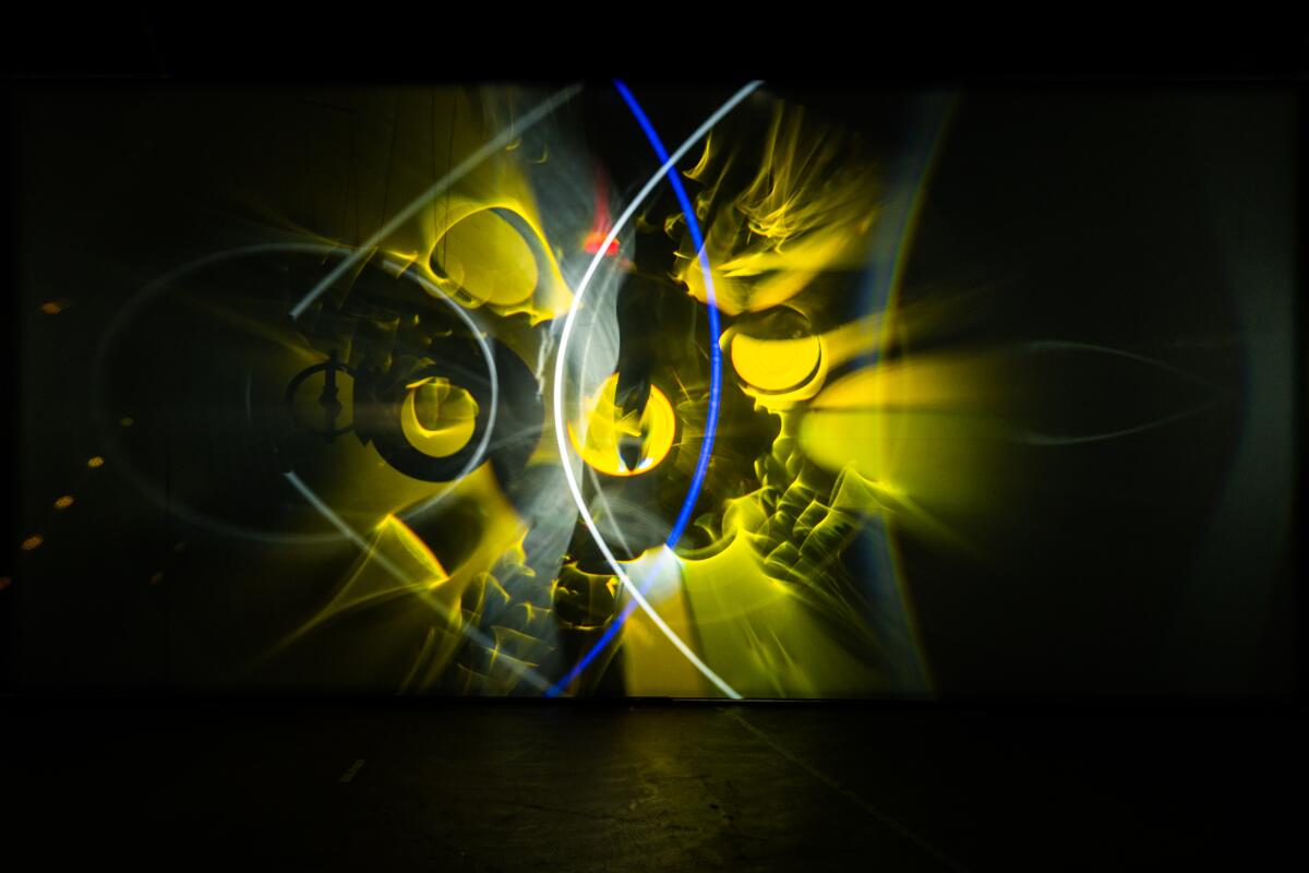 Curious abstract shapes in yellow appear on a black scrim with white and blue arcs of light. 