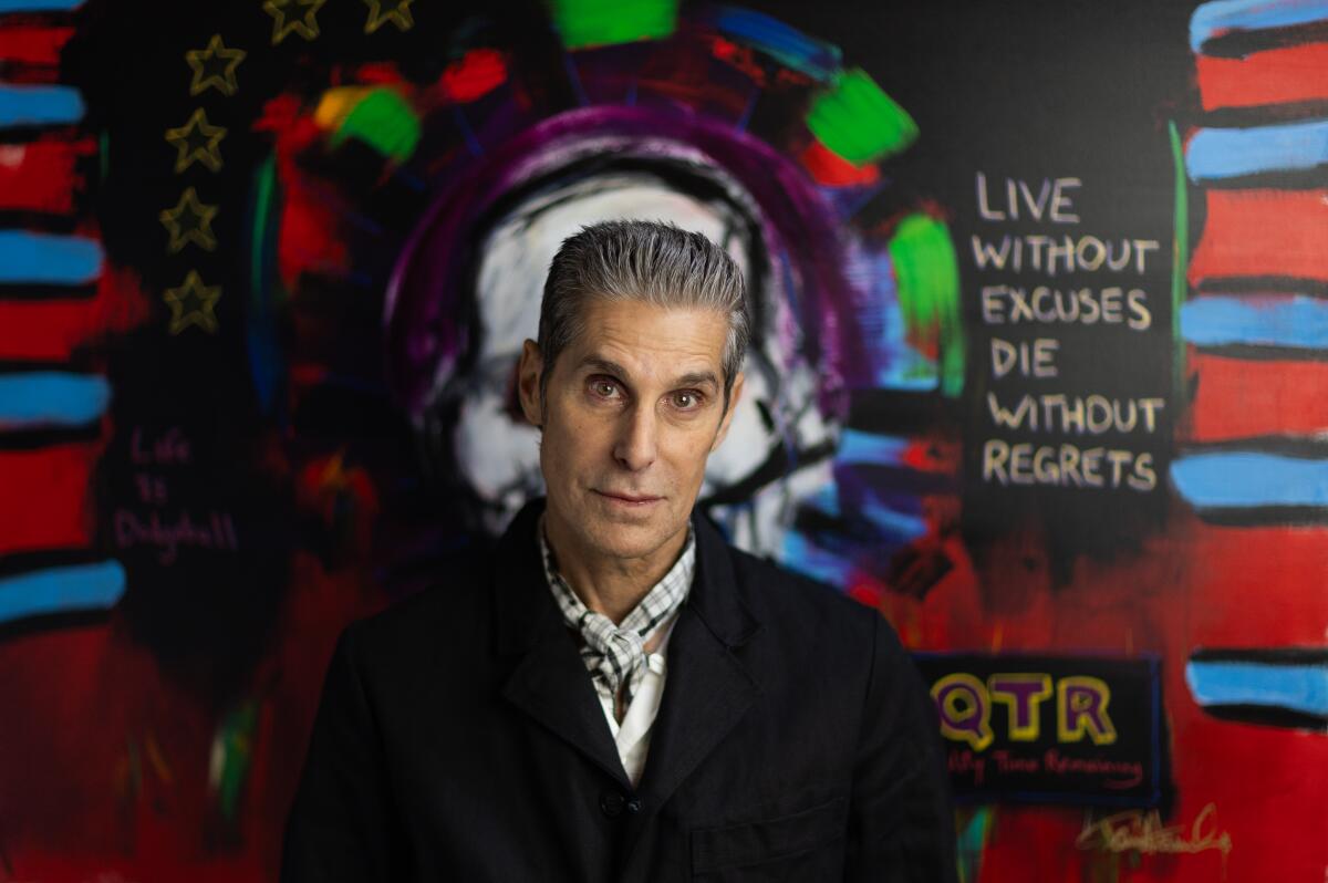 Perry Farrell in a black coat standing in front of a mural