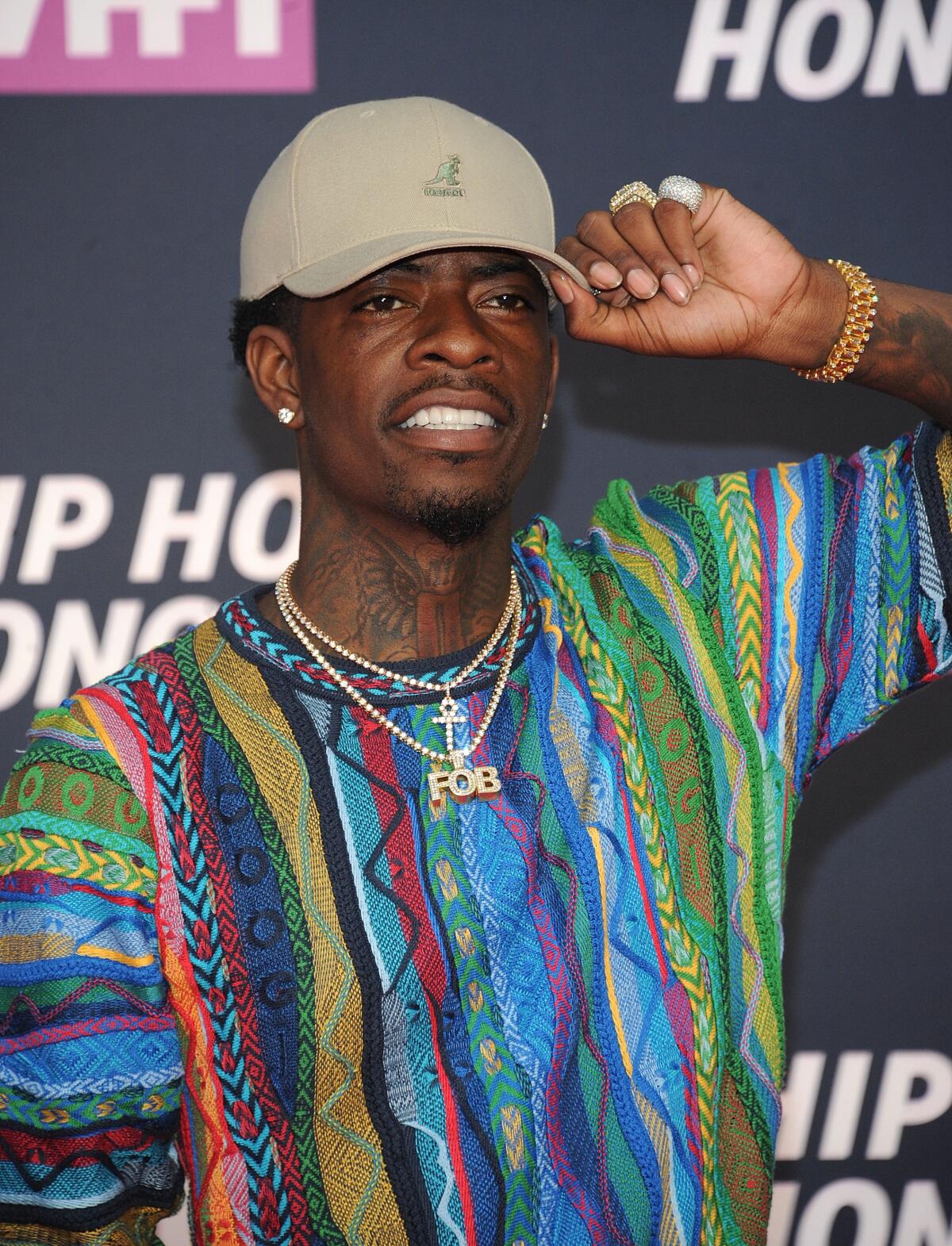 Rich Homie Quan holding the brim of a khaki hat while smiling and wearing a colorful sweater and chains