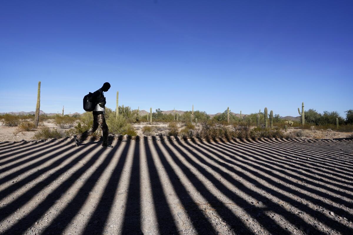 A person walks along a road shadowed by the steel columns of the border wall separating Arizona and Mexico.