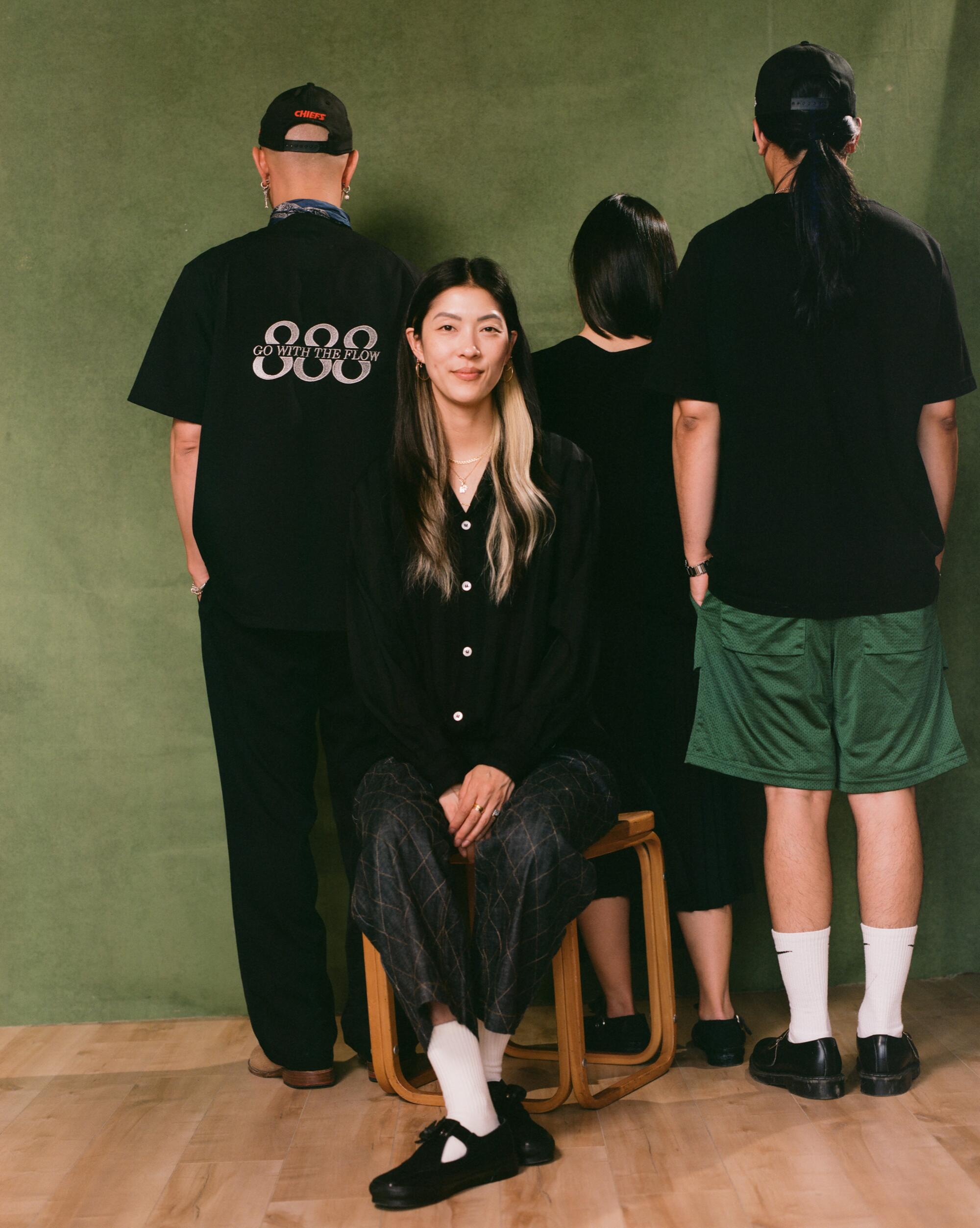 A woman sits facing forward on a stool, and three people behind her stand with their backs to the viewer.