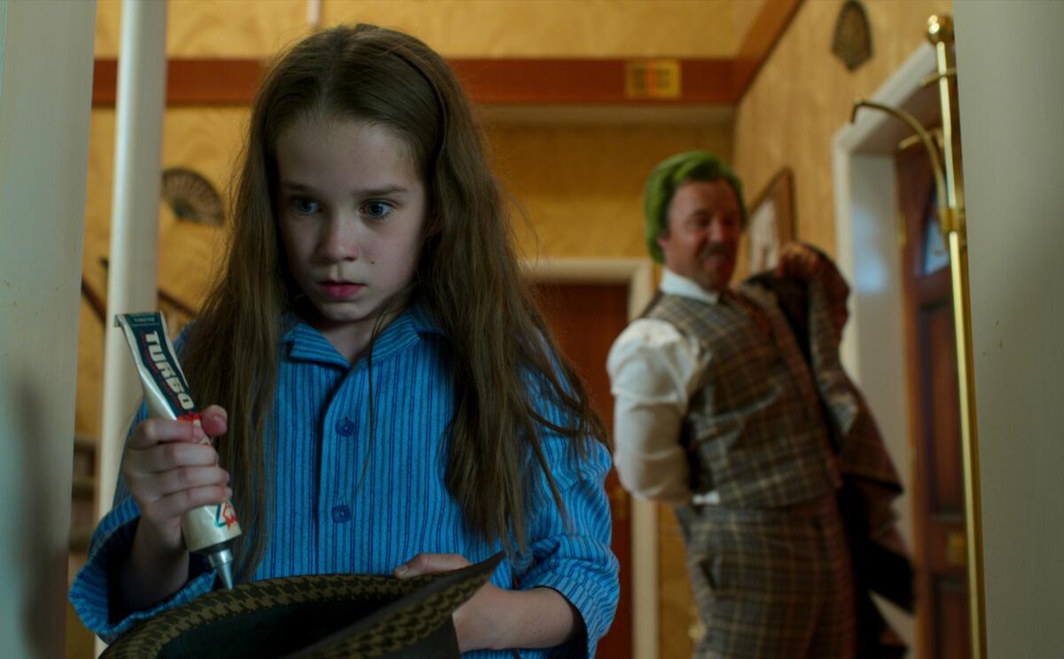 A long-haired girl squeezes a tube as a man grins behind her in the movie "Roald Dahl's Matilda the Musical."