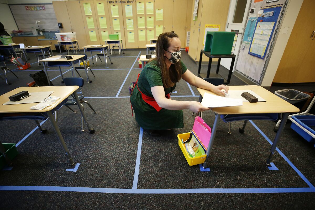 A teacher in a mask and apron kneels beside a desk as she works.