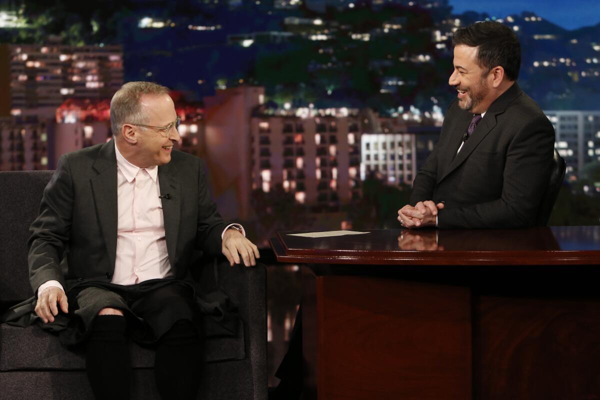 David Sedaris and Jimmy Kimmel sit and talk on the latter's show, with the Hollywood skyline as a backdrop.