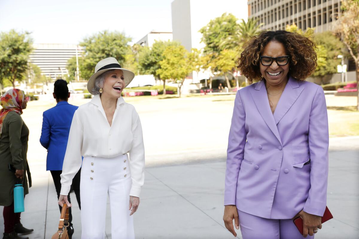A woman in a white suit and straw hat and a woman in a lavender suit laugh together.