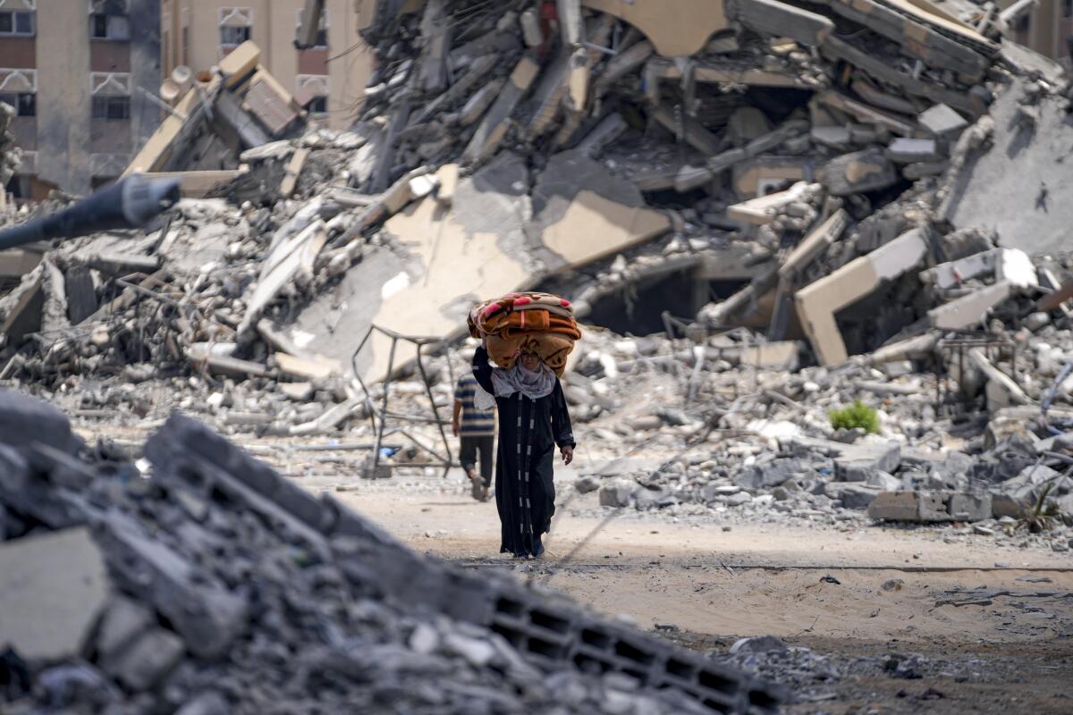 A Palestinian woman walks through massive piles of destroyed buildings, carrying belongings on her head.