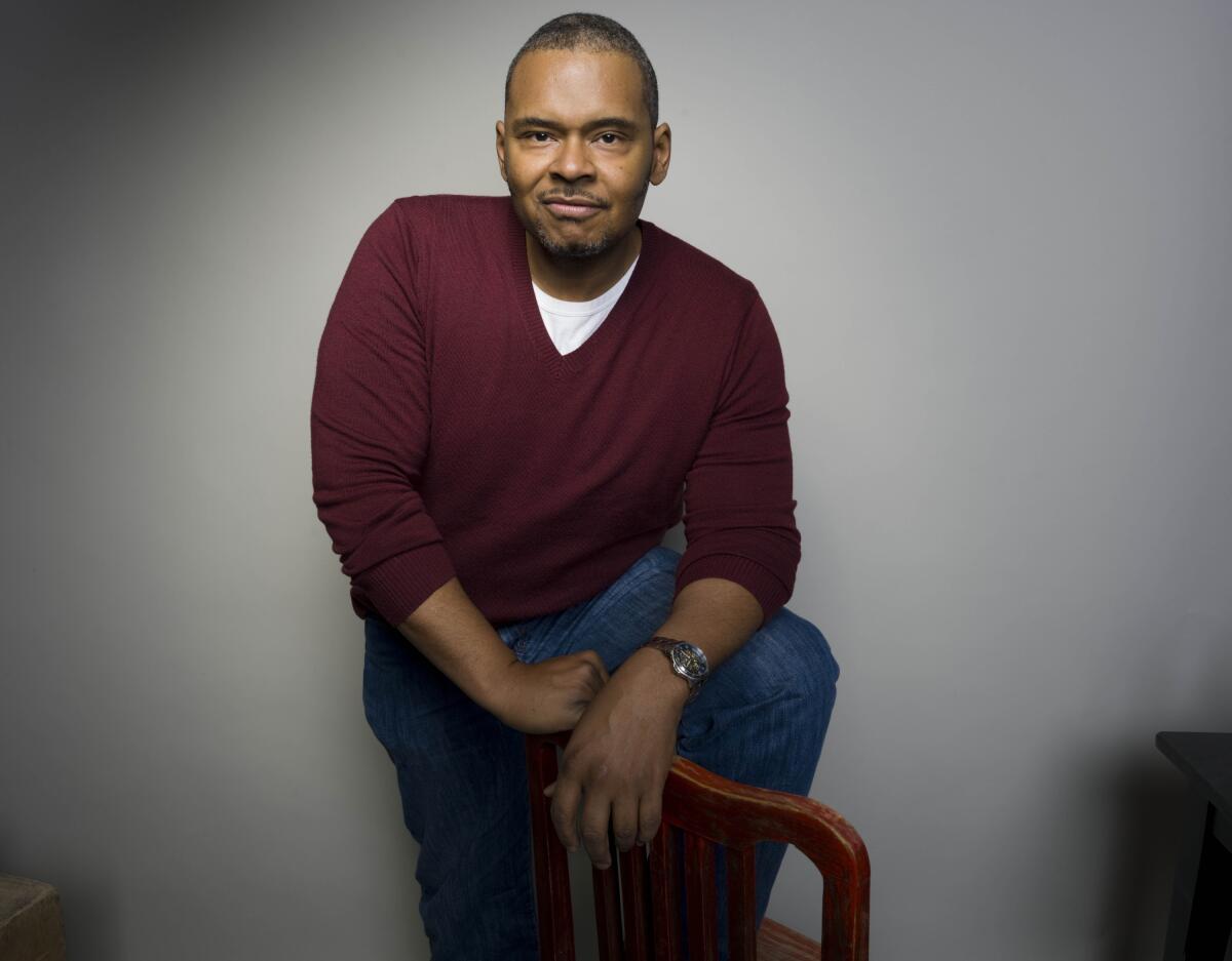 A Black man in a maroon sweater and white T-shirt and jeans posing with his left leg up on a chair against a grey background