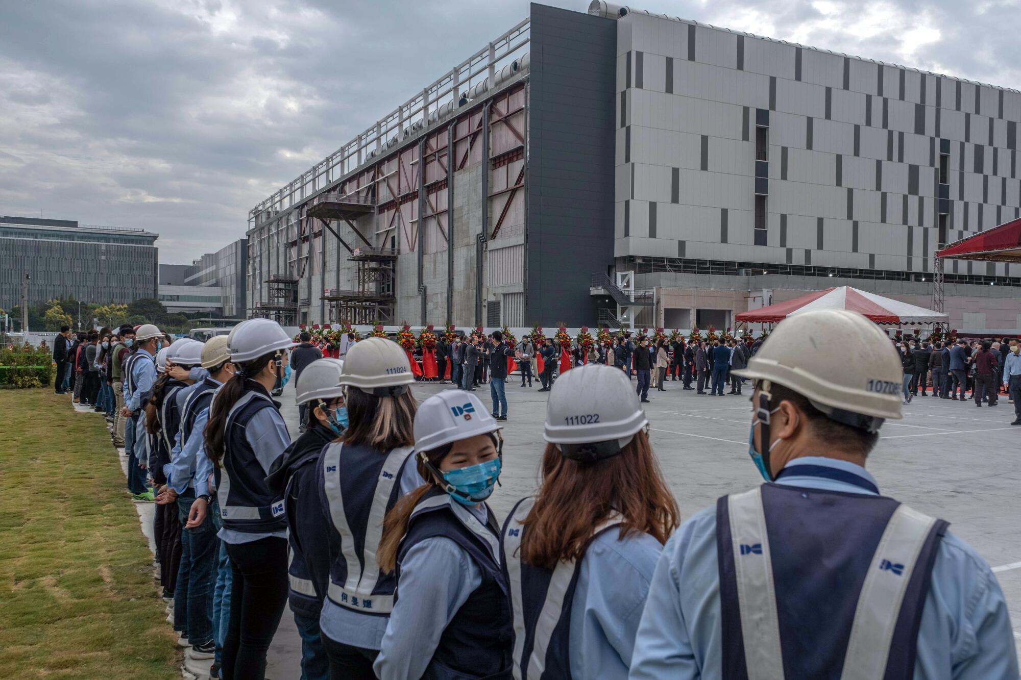 Workers in gray helmets gather for a ceremony outdoors near big buildings.