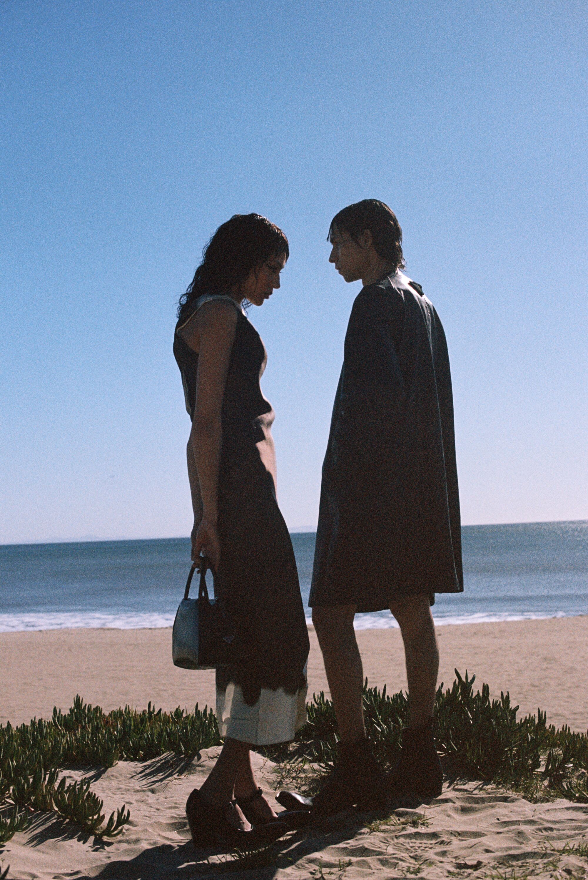 Two models on a beach, dressed in black and silhouetted by the sun.