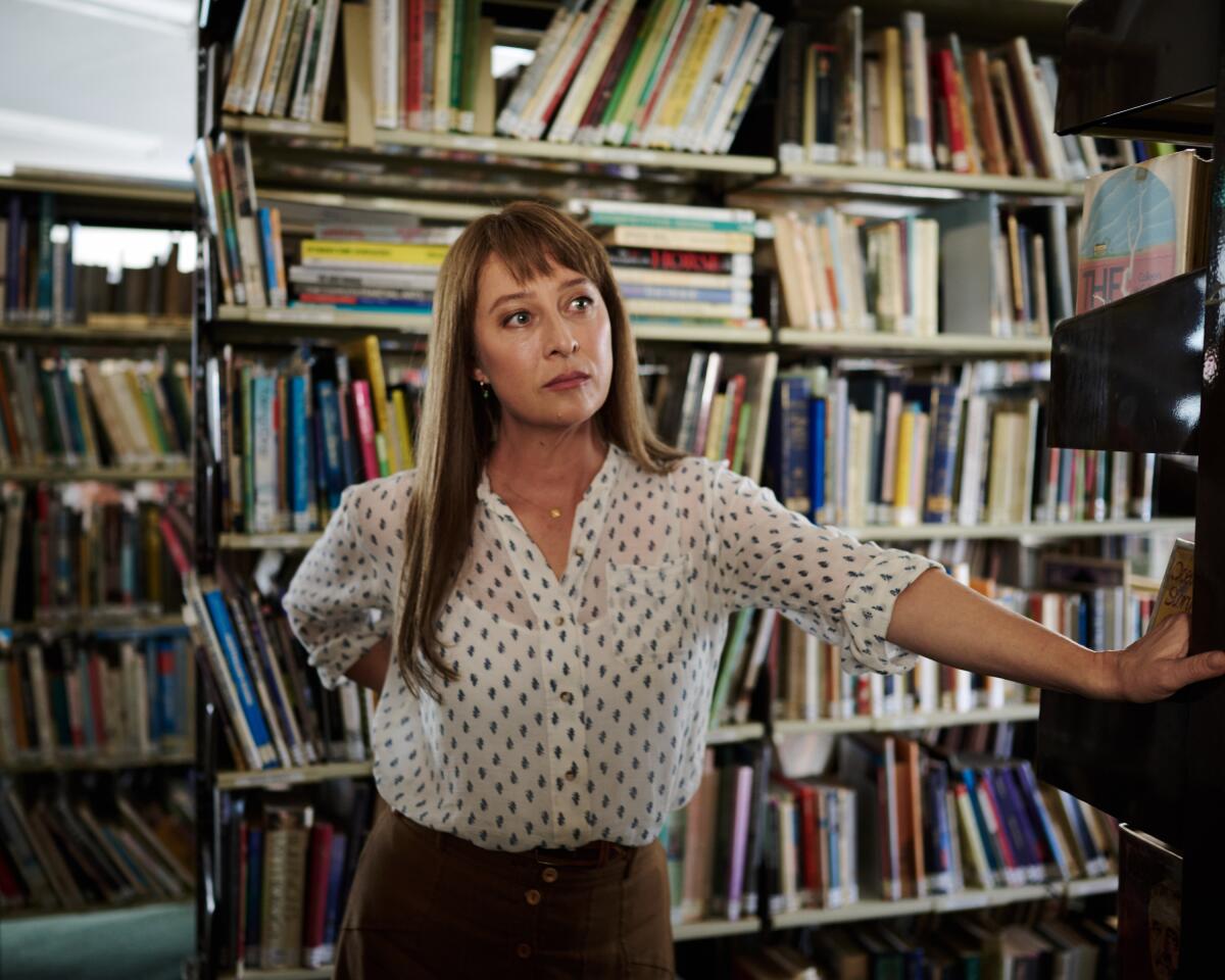 A librarian in a white shirt with polka dots leans against a bookcase. Books fill the shelves behind her.