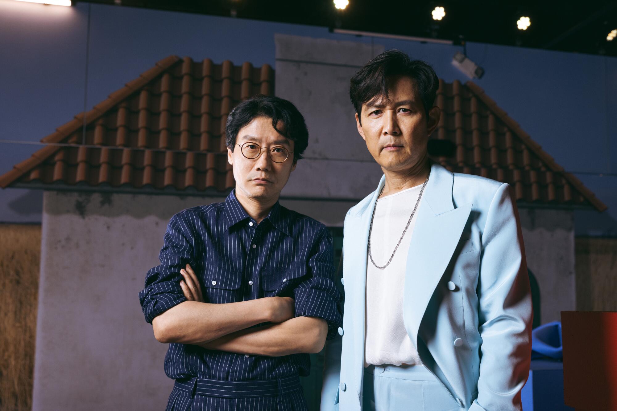 A man in a dark blue shirt folds his arms across his chest and stands next to a man in a light blue suit.