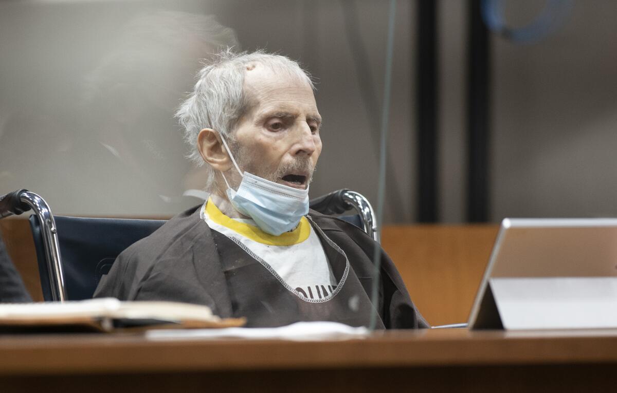 An elderly man sits in a courtroom with a facemask under his chin.