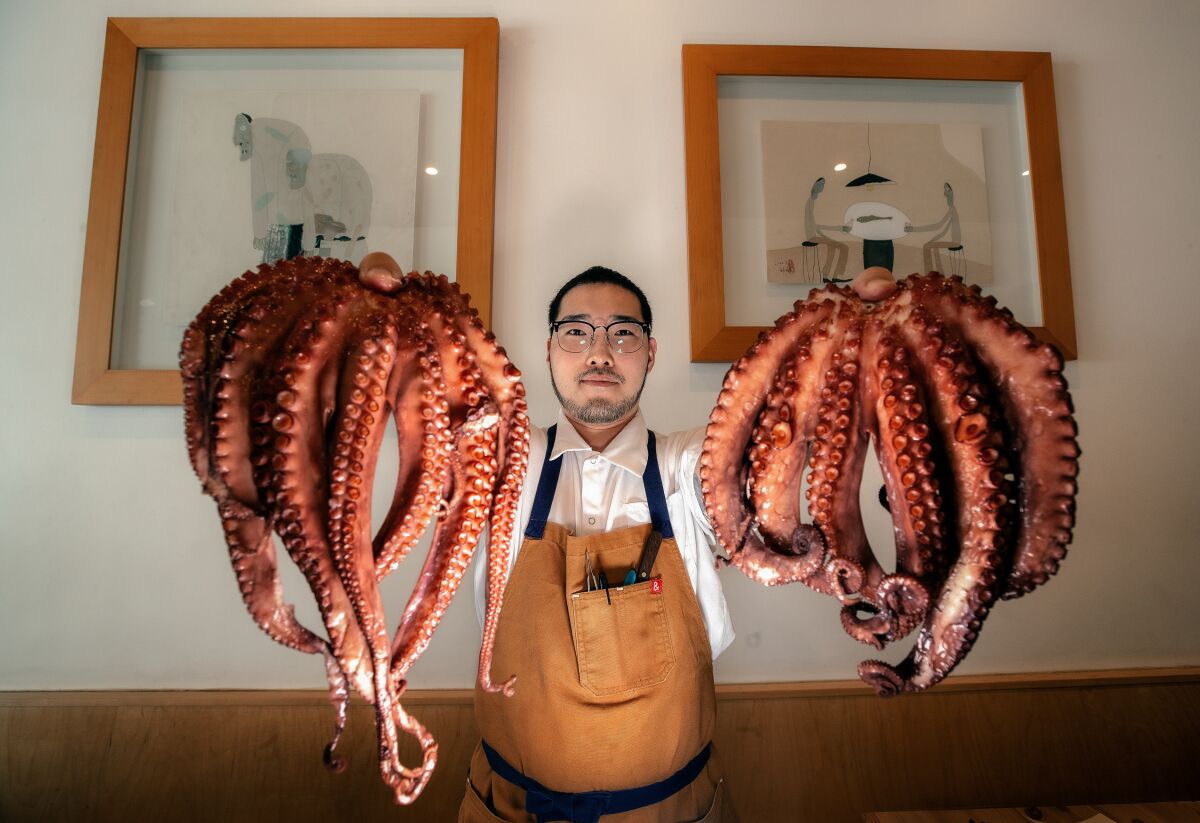 An aproned man holds up an orange-ish octopus in each hand inside a restaurant.