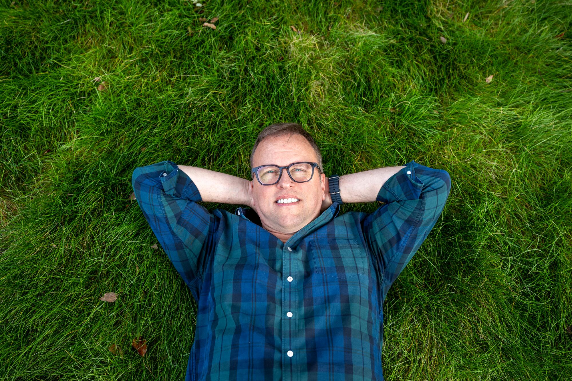A top-down photo of a man lying with his hands behind his head atop a grassy lawn.