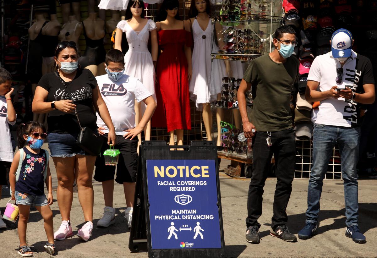 People wearing masks walk in an outdoor shopping area with a sign that says facial coverings are required