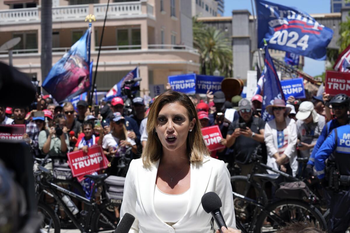 A woman speaking into several media microphones as a crowd of people raise blue and red Trump signs in the background.