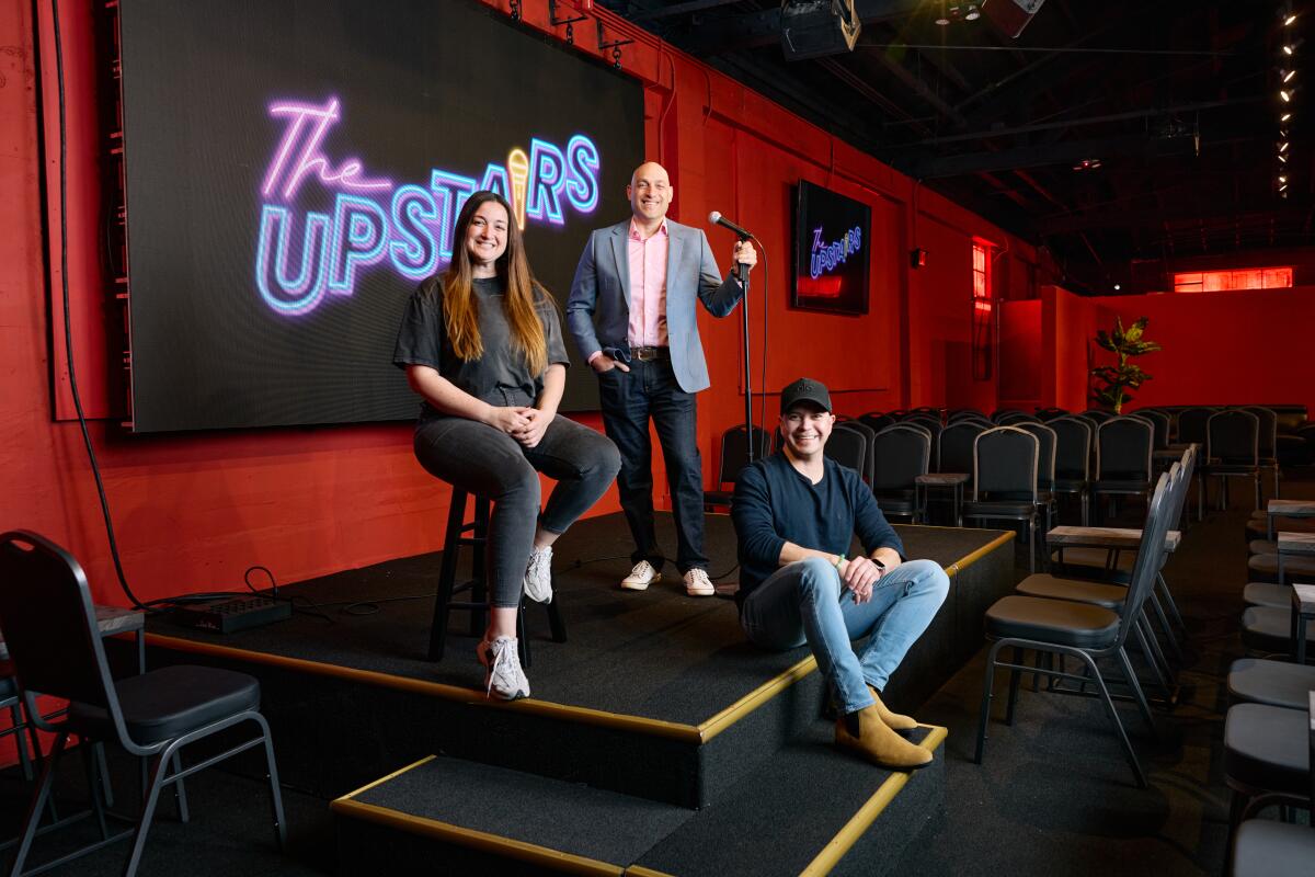 Three people on a stage with the Upstairs Comedy Club logo behind them