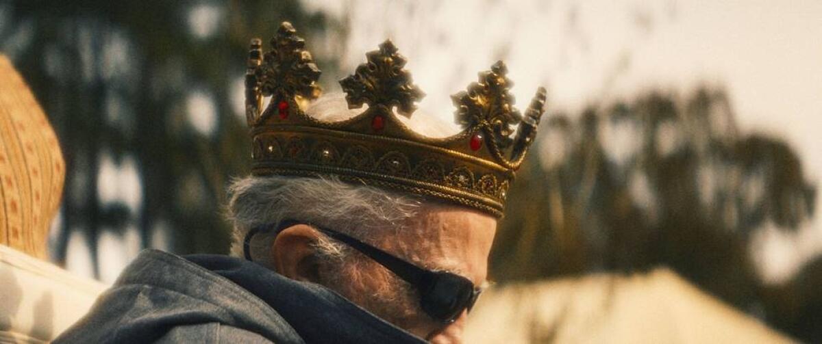 A gray haired man wears a crown and wrap-around dark sunglasses.