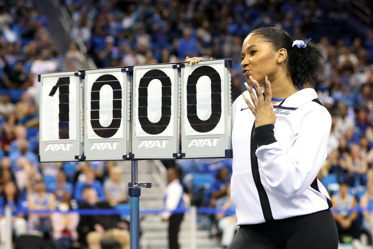 A woman in a white warmup top and black pants holds up her hand and strikes a pose next to a sign displaying 10.00.