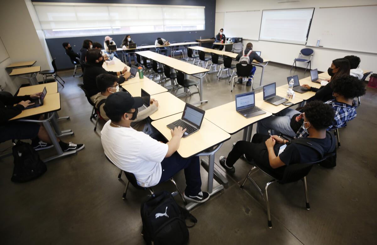 Masked students, many in close proximity, sit at desks arranged in a U shape 